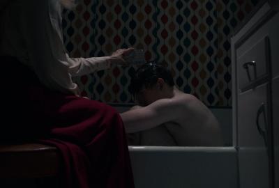 A woman pours water over a shirtless young man curled up in a bathtub in a dim, retro-patterned bathroom.